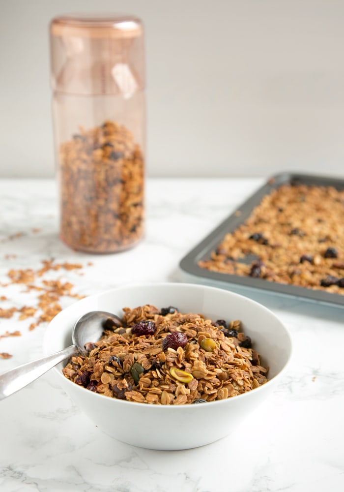 Christmas Homemade Granola in a small bowl with a spoon, more granola in a container and on a tray in the background.