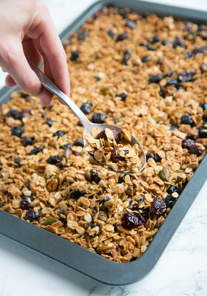 Hand holding a spoon filled with Christmas homemade granola, over a tray filled with more granola.