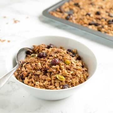 Christmas Homemade Granola in a small bowl with a spoon, more granola on a tray in the background.