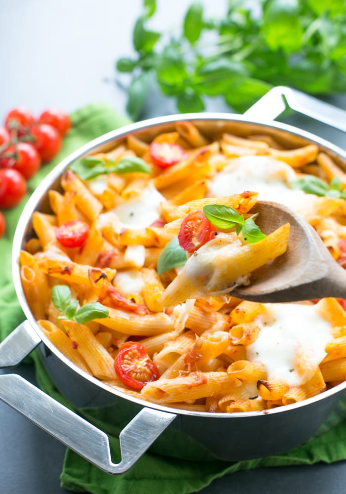 wooden spoon holding cheesy pasta over a pan filled with caprese pasta bake