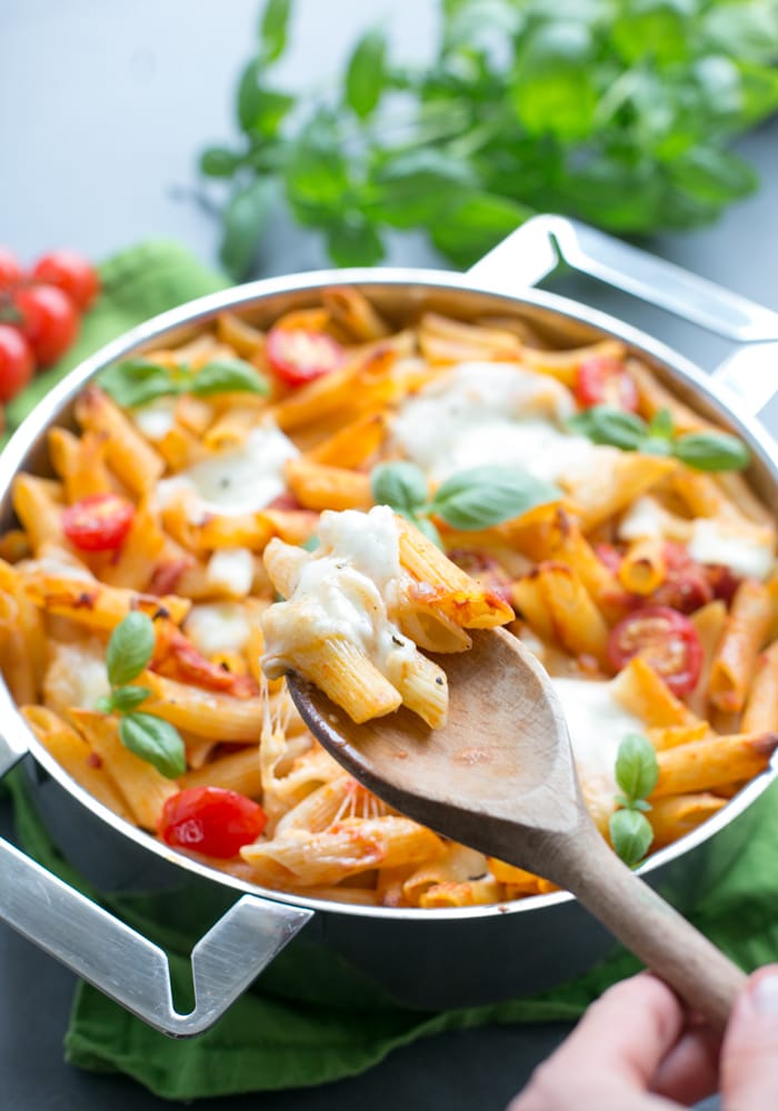 wooden spoon holding cheesy pasta over a pan filled with caprese pasta bake