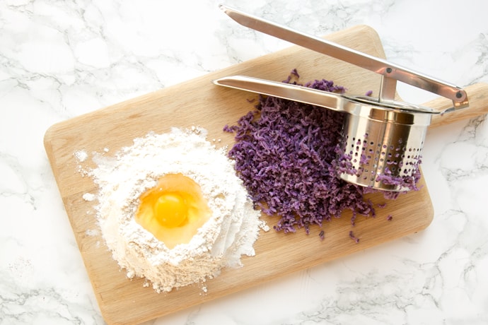 egg cracked in a small mountain of flour, next to potato ricer and purple potato mixture.