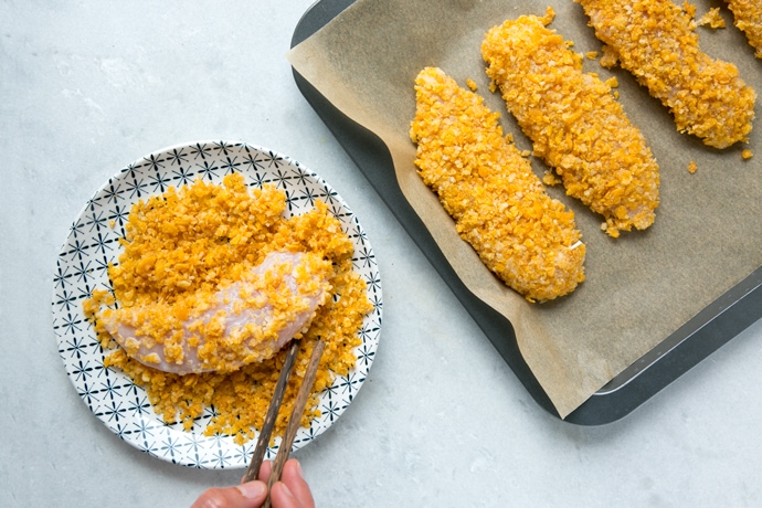 chicken breas being coated in coating mix breadcrumbs, next to a tray of already coated chicken breasts.