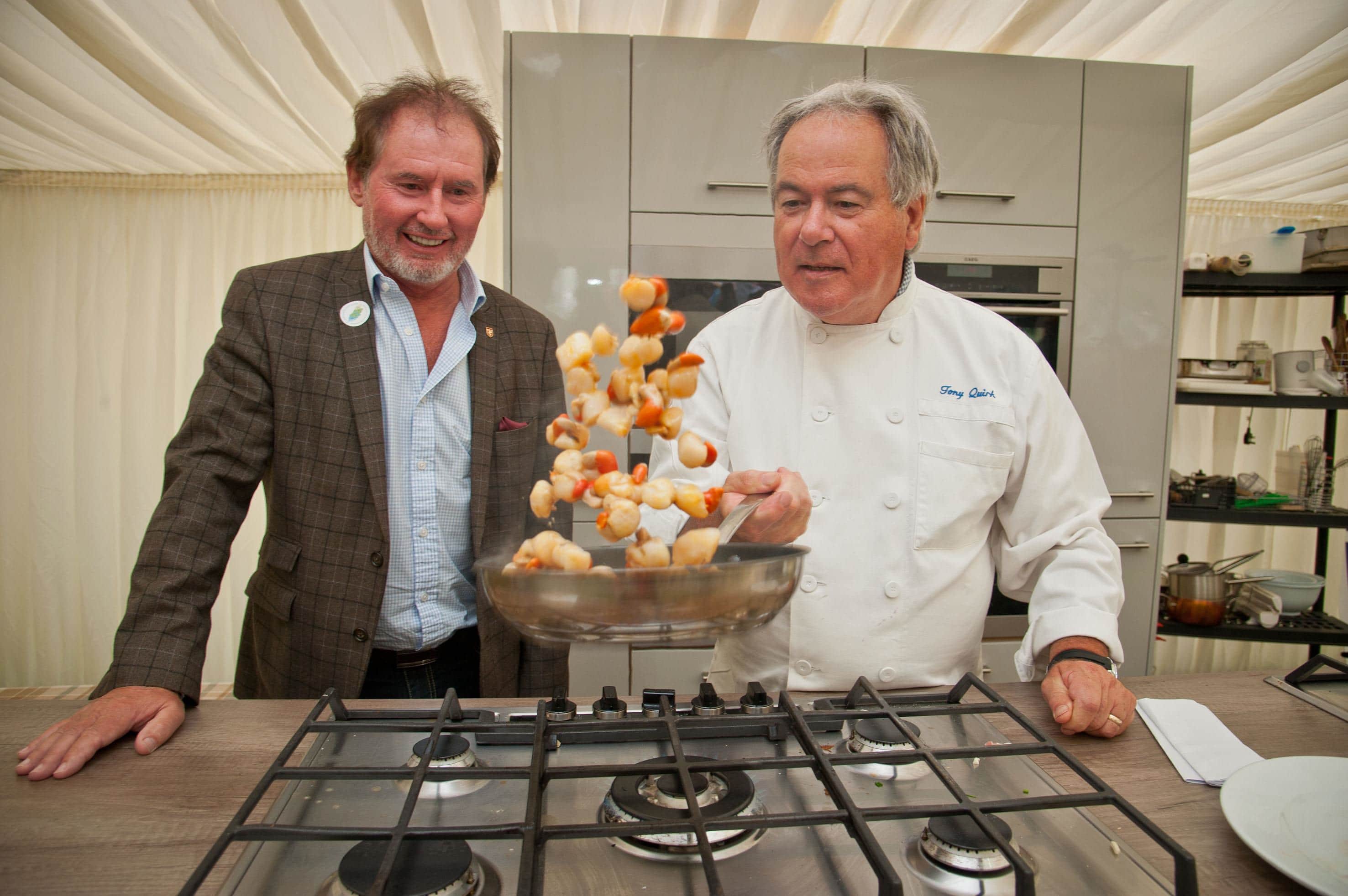 Chef Tony Quirk cooking queenies in a pan over the hobs, next to Hon Geoffrey Boot, Minister for Environment, Food and Agriculture.