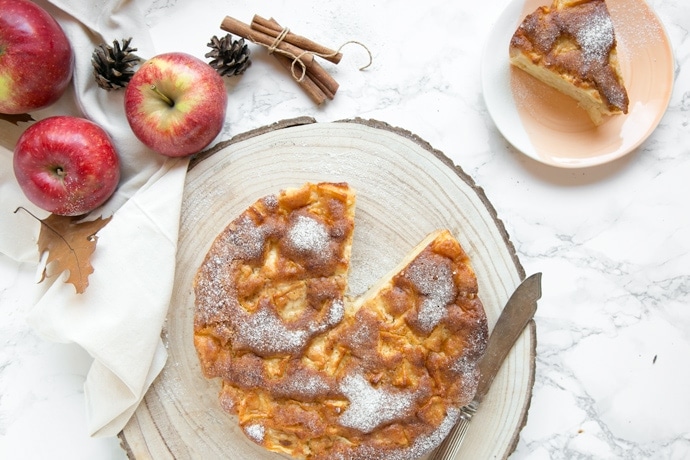 Italian apple cake on wood board next to apples, cinnamon sticks and a small plate with a slice of cake