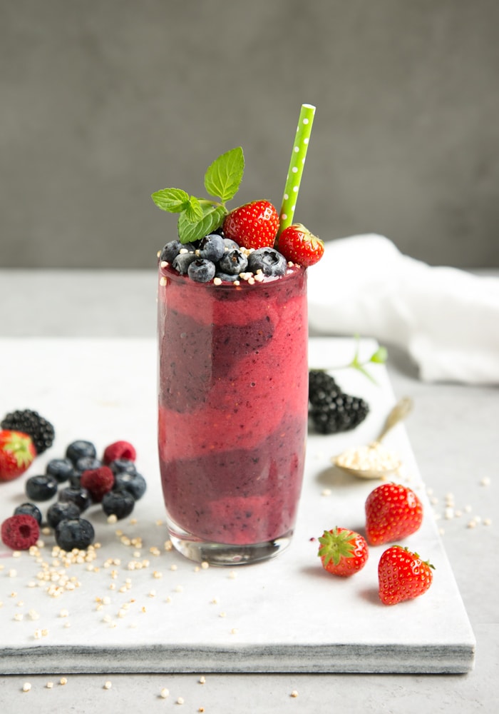 mixed berry smoothie in tall glass decorated with berries, mint and green stroll, on white marble board and berries and a spoon with puffed quinoa for decoration