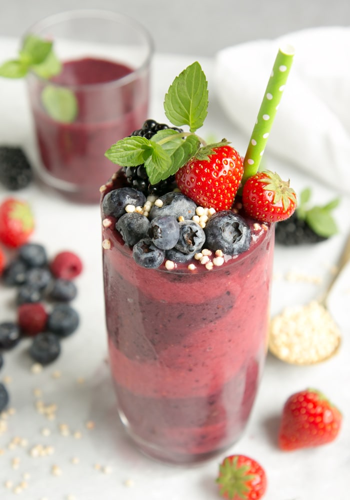 mixed berry smoothie in tall glass decorated with berries, mint and green stroll, white background with glass filled with smoothie , berries and a spoon with puffed quinoa