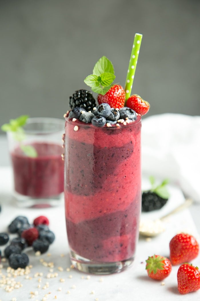 mixed berry smoothie in tall glass decorated with berries, mint and green stroll, on white marble board with glass fillec with smoothie and berries and a spoon with puffed quinoa for decoration