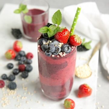 mixed berry smoothie in tall glass decorated with berries, mint and green stroll, on white marble board with smoothie in another glass in the background and berries and a spoon with puffed quinoa for decoration
