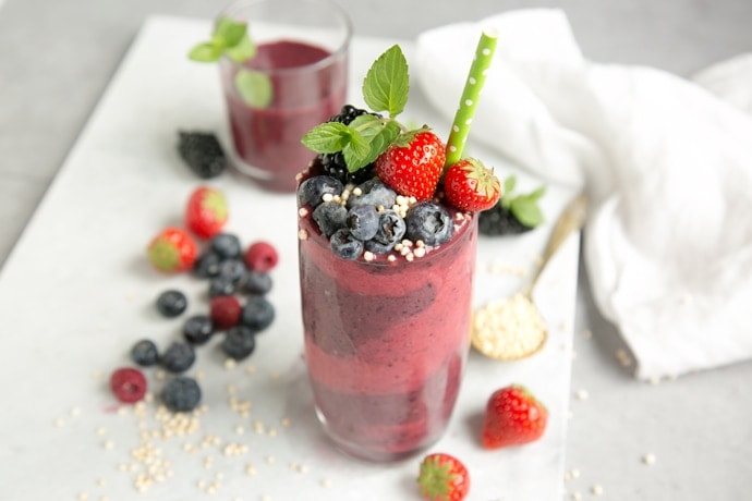 mixed berry smoothie in tall glass decorated with berries, mint and green stroll, on white marble board with smoothie in another glass in the background and berries and a spoon with puffed quinoa for decoration