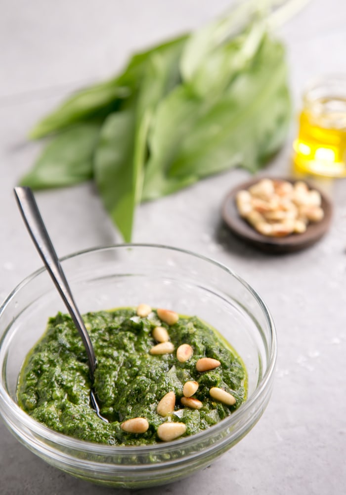 wild garlic pesto in small glass bowl, with pine nuts, wild garlic and olive oil in the background