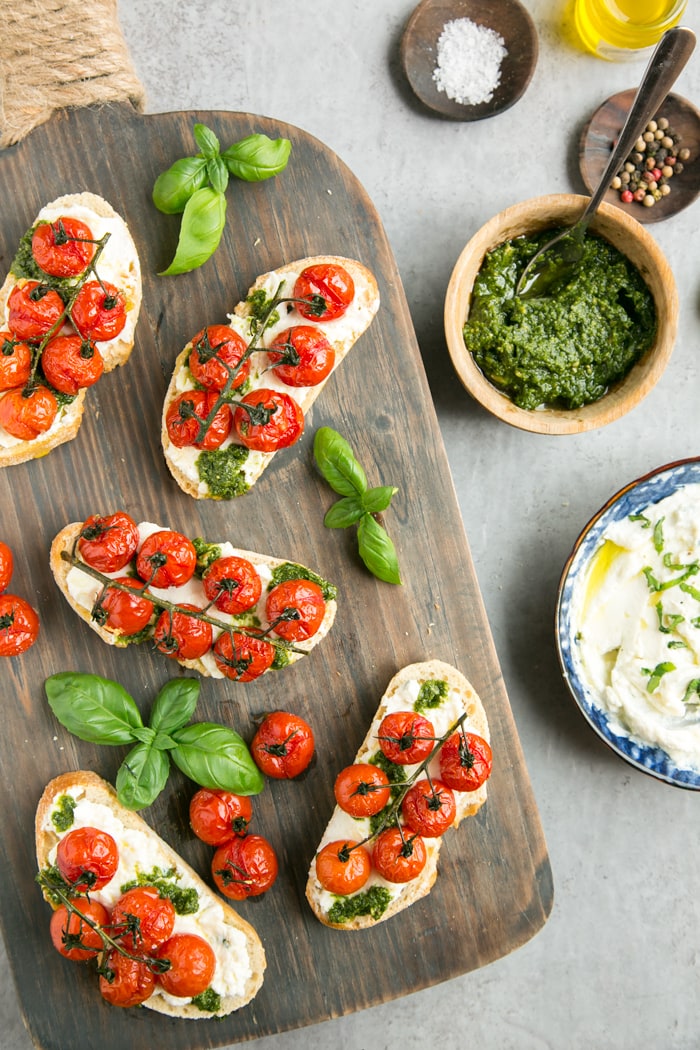 confit tomato bruschettas with ricotta and basil pesto on a wood board next to bowl of basil pesto, and bowl with ricotta cheese