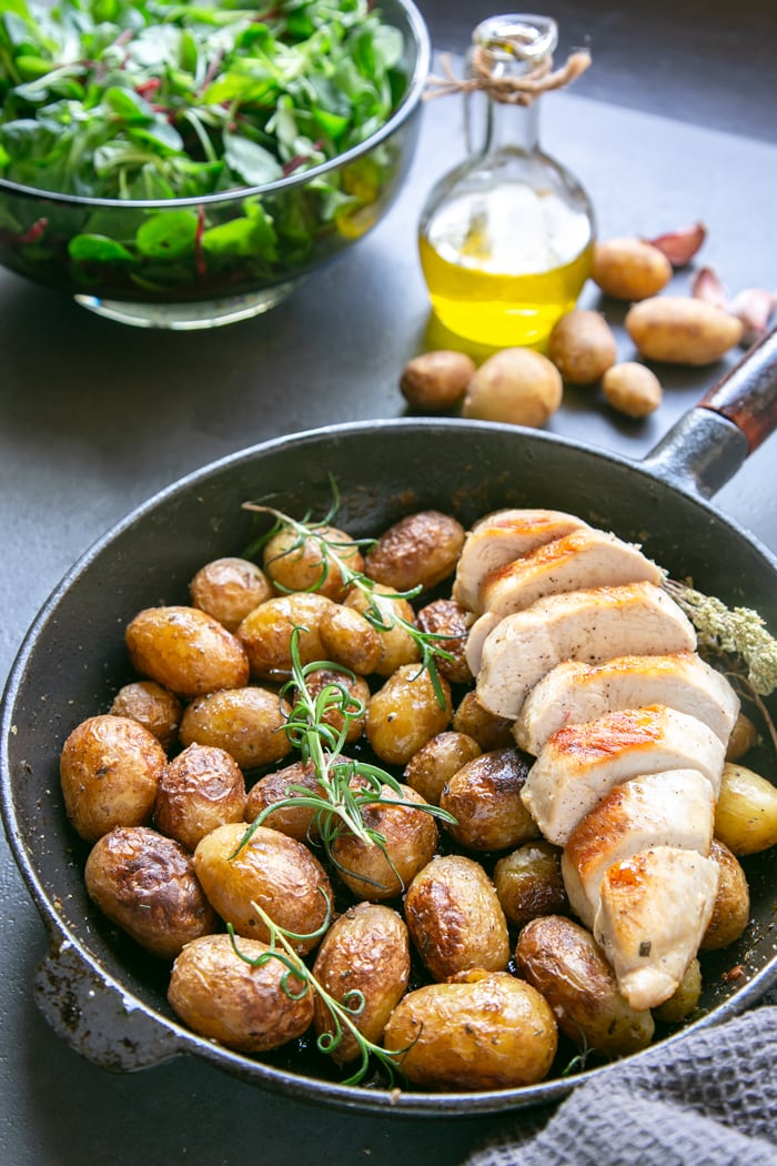 Italian pan roasted new potatoes with rosemary in skillet with grilled chicken breast, olive oil bottle, salad in bowl and raw potatoes in the background