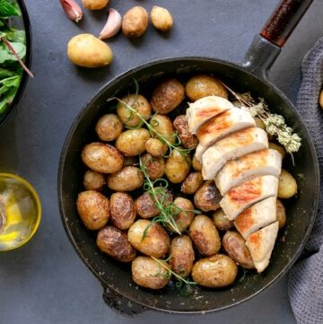 Italian pan=roasted new potatoes in skillet with grilled chicken breast, salad bowl and olive oil bottle on the left side, potatoes arranged on the background and grey towl on the right side