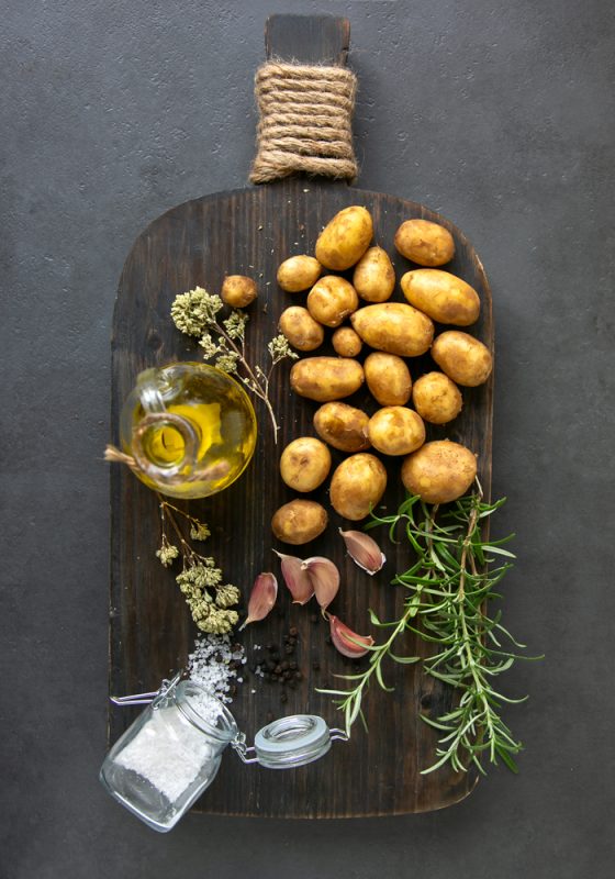 recipe ingredients: potatoes, olive oil bottle, rosemary sprigs, garlic, oregano sprigs and sea salt on a chopping board