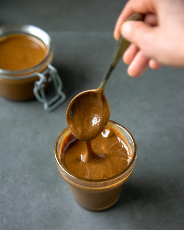 homemade dulce de leche in small glass jar with hand holding a teaspoon dripping some dulce de leche