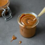 homemade dulce de leche in small glass jar with teaspon inside, jar of dulce de leche in the background