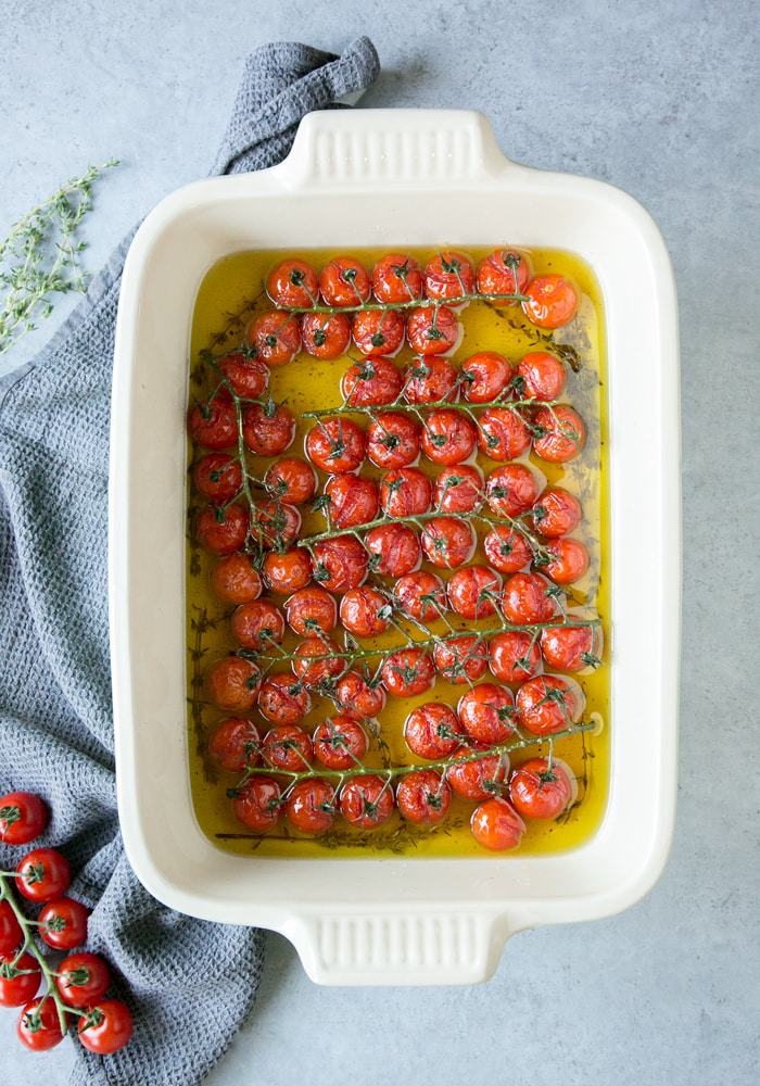 tomato confit in a large white baking dish, grey cloth on the side and fresh cherry tomatoes on the vine