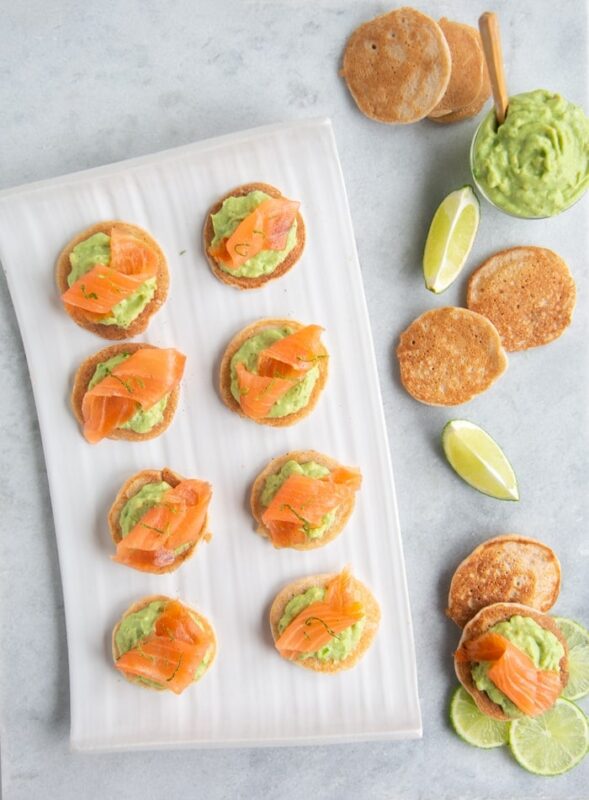 gluten-free blini with avocado cream and smoked salmon on white plate next to avocado cream in bowl, plain blini and slices of lime