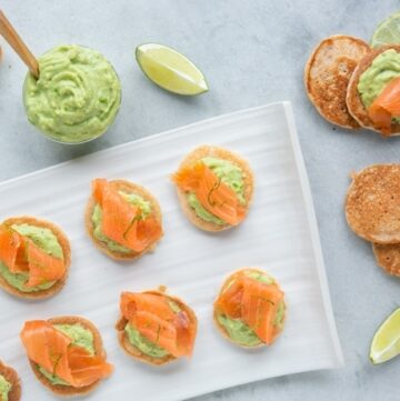 gluten-free blini topped with avocado cream and salmon, served with lime wedges and extra avocado cream on the side