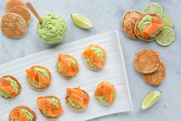 gluten-free blini topped with avocado cream and salmon, served with lime wedges and extra avocado cream on the side
