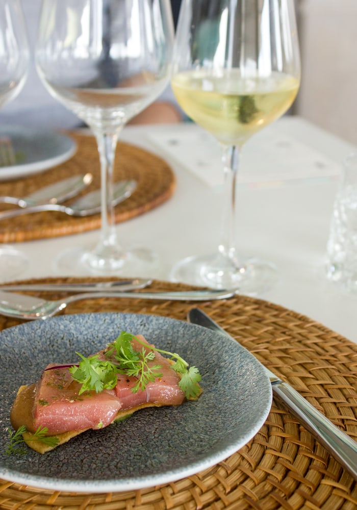 fresh tuna fillet on toast and charro black beansand guacamole, served on a blue plate with white wine on the side, table setting in the background