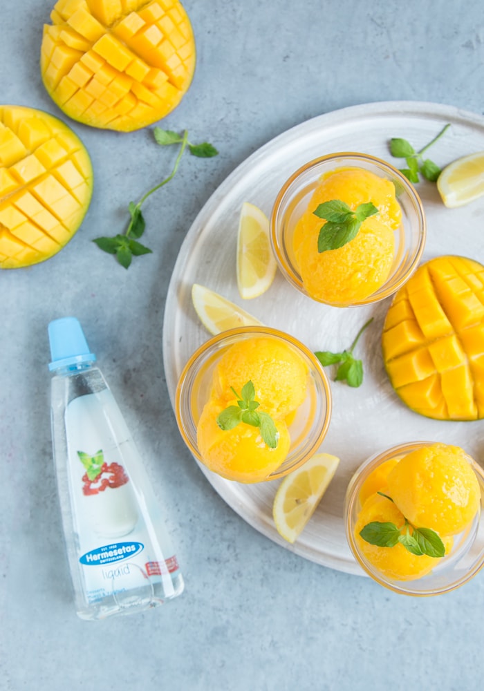 vegan mango sorbet topped with mint leaves in two glasses, with lemon wedges and mango, and Hermesetas Liquid sweetener in the background 