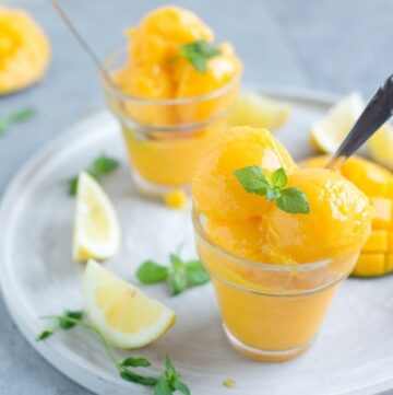 vegan mango sorbet topped with mint leaves in two glasses, with lemon wedges and mango in the background