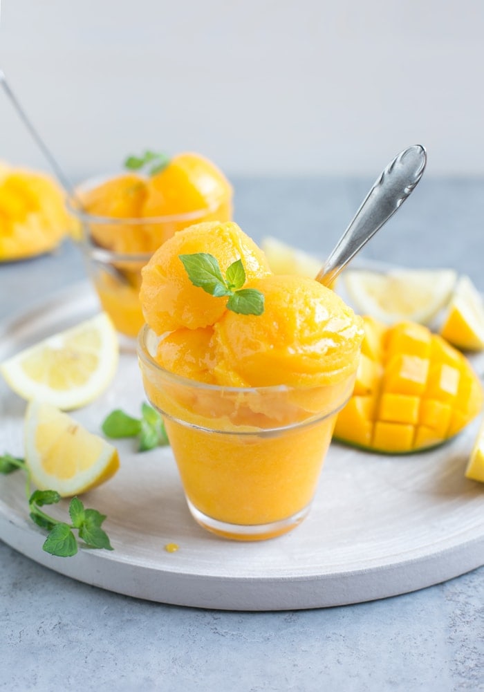 vegan mango sorbet topped with mint leaves in two glasses, with lemon wedges and mango in the background