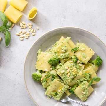 Broccoli pesto pasta on grey plate, raw broccoli, lemon, basil leaves, pine nuts and paccheri pasta in the background