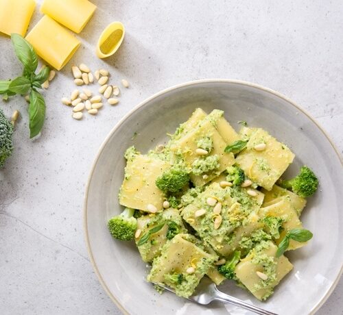 Broccoli pesto pasta on grey plate, raw broccoli, lemon, basil leaves, pine nuts and paccheri pasta in the background