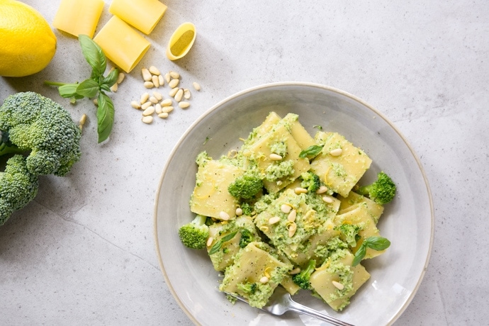 Broccoli pesto pasta on grey plate, raw broccoli, lemon, basil leaves, pine nuts and paccheri pasta in the background