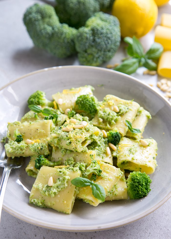 Broccoli pasta on grey plate, raw broccoli, lemon, basil leaves, pine nuts and paccheri pasta in the background