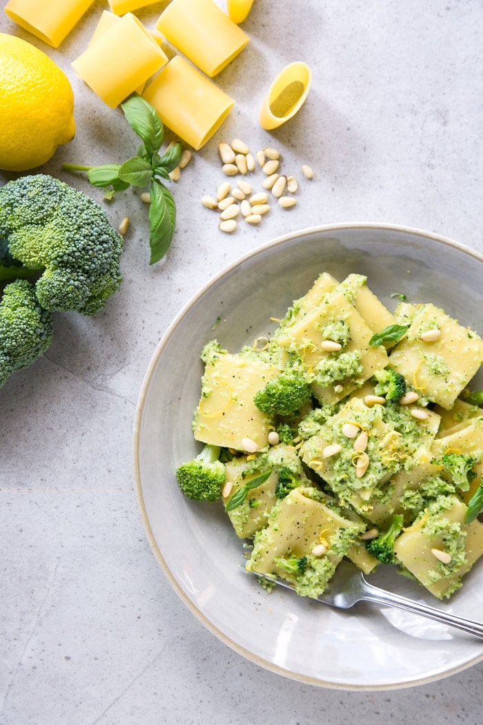 Broccoli pesto pasta on grey plate, raw broccoli, lemon, basil leaves, pine nuts and paccheri pasta in the background