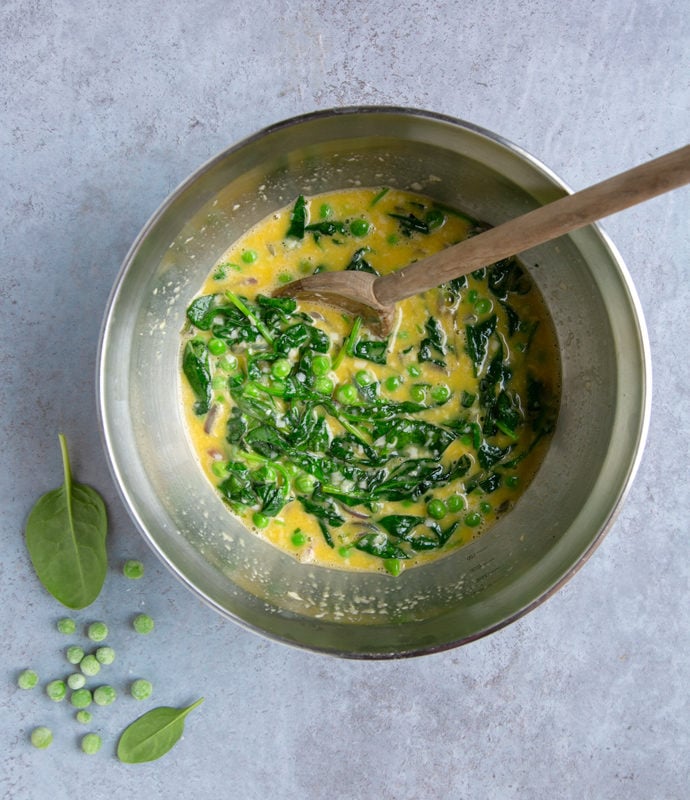 italian frittata with spinach and peas preparation step 3: egg mixture, cooked spinach and peas in large metal bowl and wooden spoon, spinach leaves and frozen peas next to the bowl for decoration