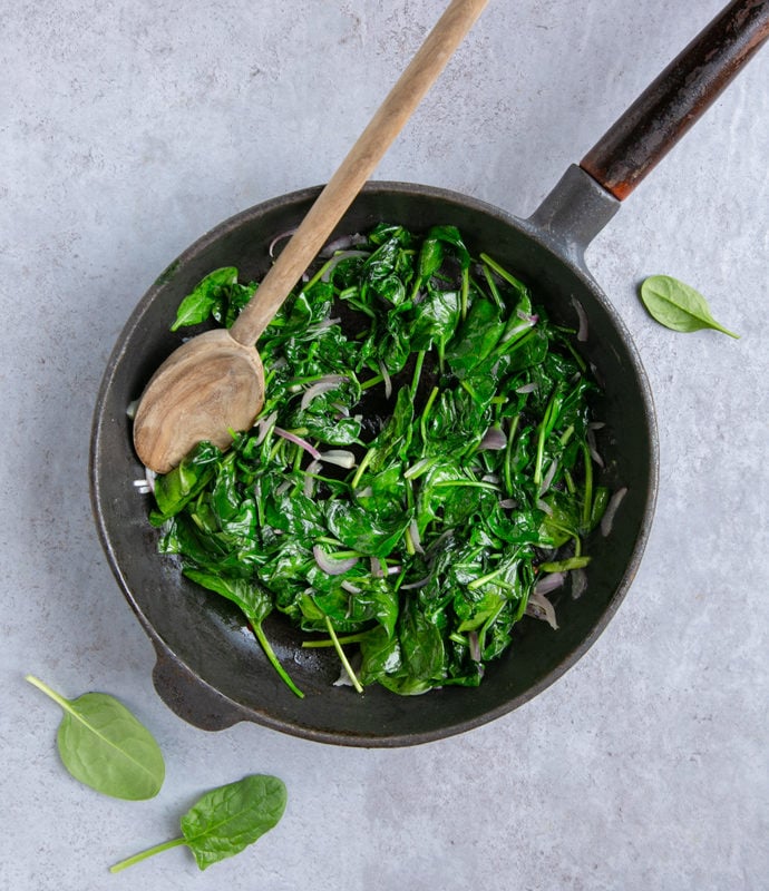 italian frittata with spinach and peas preparation step 1: cooked spinach and onion in a cast iron skillet, wood spoon on top, spinach leaves and peas next to the pan for decoration