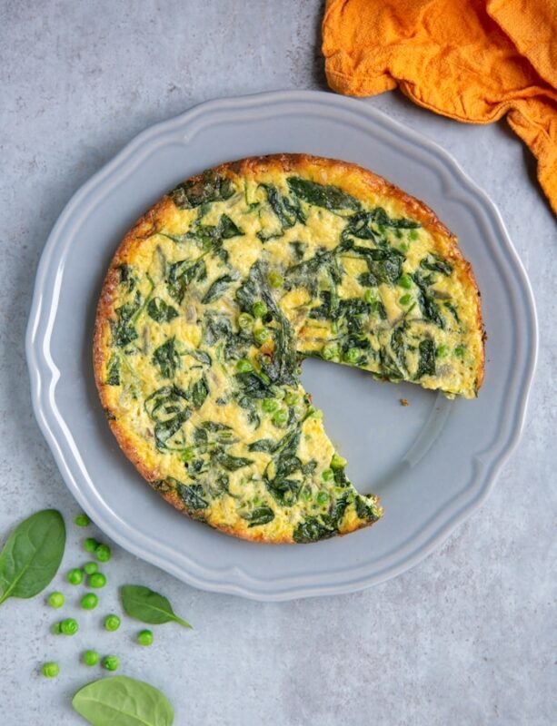 italian frittata with spinach and peas with one wedge removed on grey plate, next to orange cloth, and spinach leaves and peas for decoration