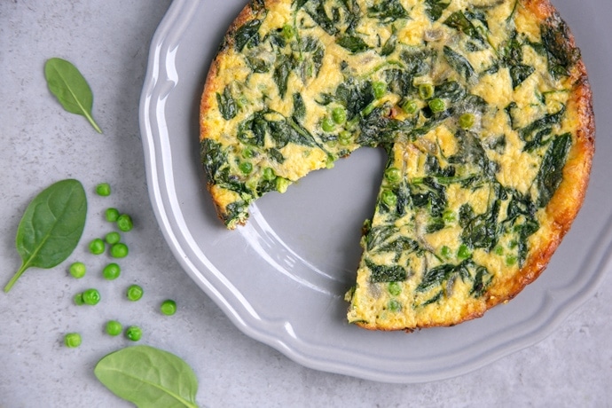 italian frittata with spinach and peas with one wedge removed on a grey plate, spinach leaves and peas on the side for decoration