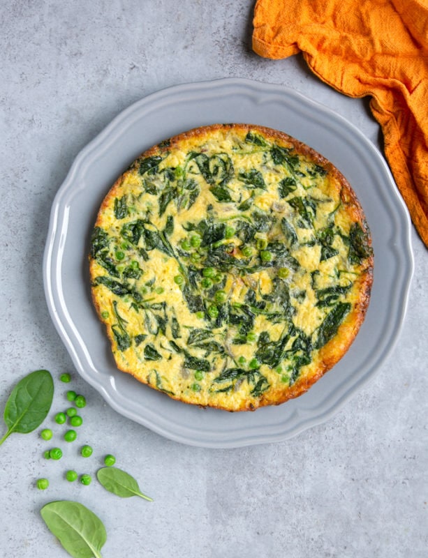 italian frittata with spinach and peas on large grey plate, next to orange cloth, and spinach leaves and peas for decoration