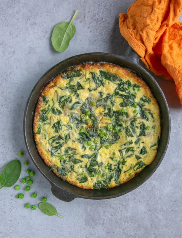 italian frittata with spinach and peas preparation step 5: cooked frittata in cast iron skillet, orange cloth covering the handle, spinach leaves and peas next to the skillet for decoration