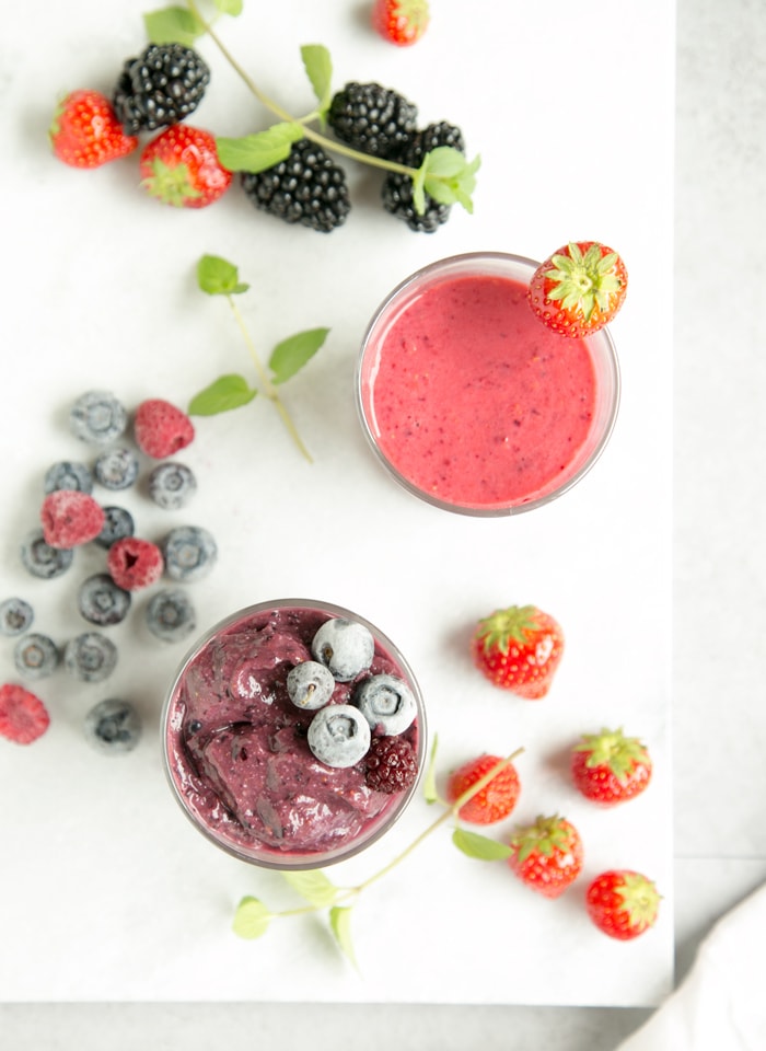 glass with blackberry and blueberry smoothie next to a strawberry and raspberry smoothie, mixed berries in the background over a white marble board