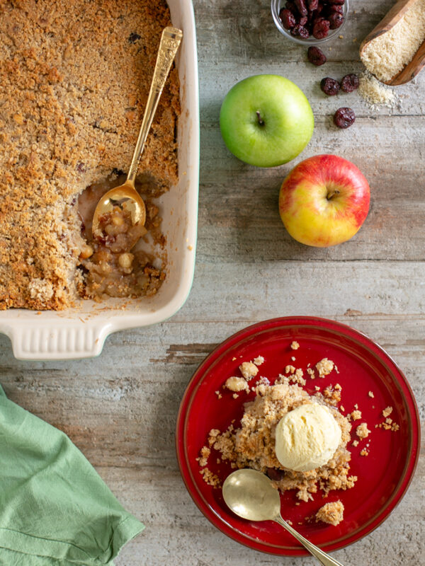 On the right down side, vegan apple crumble on a red plate topped with vanilla ice cream and a spoon next to the plate. On the top right side 2 apples, one green and one red, cranberries in a small glass pot and some almond flour scattered over the counter. On the top left side, apple crumble in a baking dish with a gold spoon.