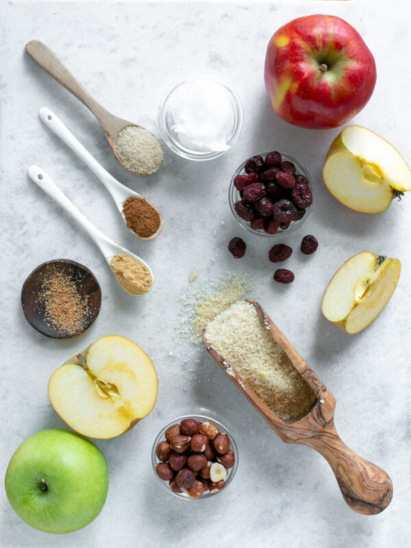 vegan apple crumble ingredients: 1 red apple, half apple plus two extra slices, one green apple, 1 teaspoon of nutmeg, one teaspoon of ginger powder, one teaspoon of cinnamon, 1 tablespoon of sugar, one small pot with coconut oil, one small pot with cranberries, one small pot with toasted hazelnuts, and 1 flour spoon with almond flour.