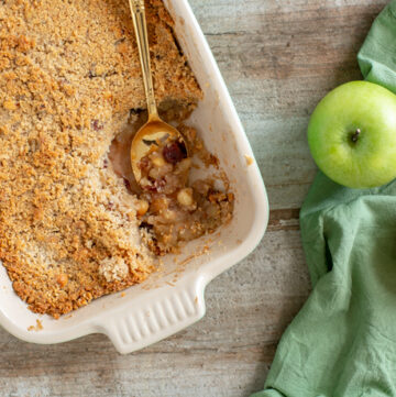 vegan apple crumble in a baking dish with a golden spoon, one red apple and one green apple on the right side over a green napkin.