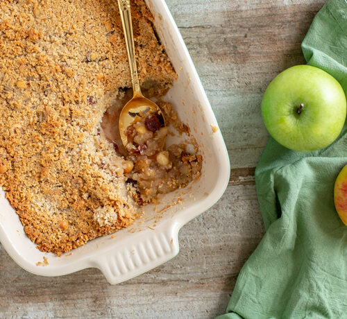 vegan apple crumble in a baking dish with a golden spoon, one red apple and one green apple on the right side over a green napkin.