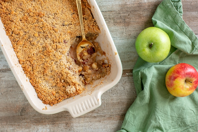 vegan apple crumble in a baking dish with a golden spoon, one red apple and one green apple on the right side over a green napkin.