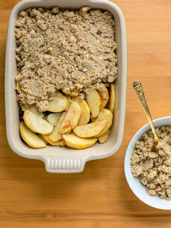 recipe step 2: baking dish with apple filling topped half-way through with the crumble topping, a small white plate filled with crumble topping and a spoon, next to the baking dish