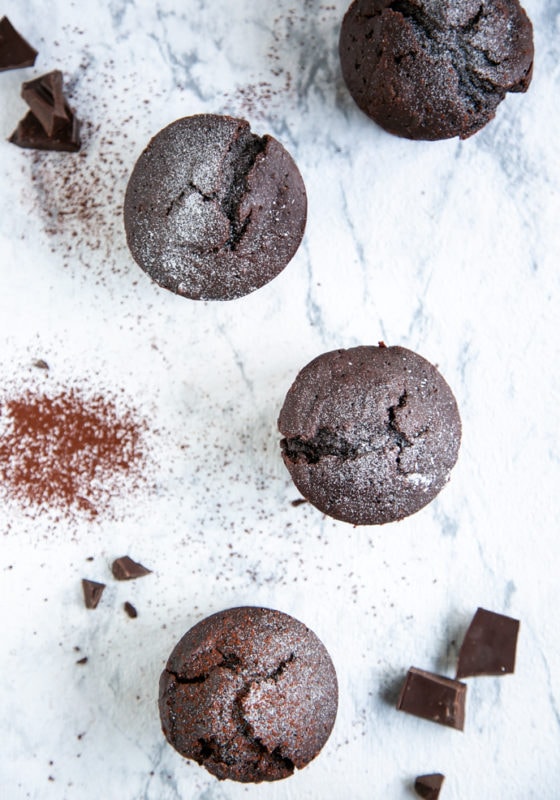 four vegan chocolate muffins over white marble background, cocoa powder and chocolate chunks sprinkled over