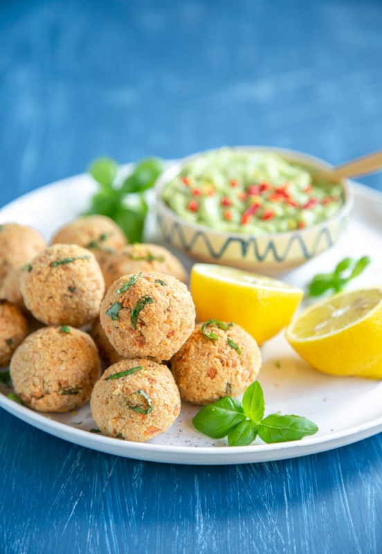 baked salmon meatballs on white plate served with halved lemon, basil leaves and avocado cream in a small pot, can of salmon in the background