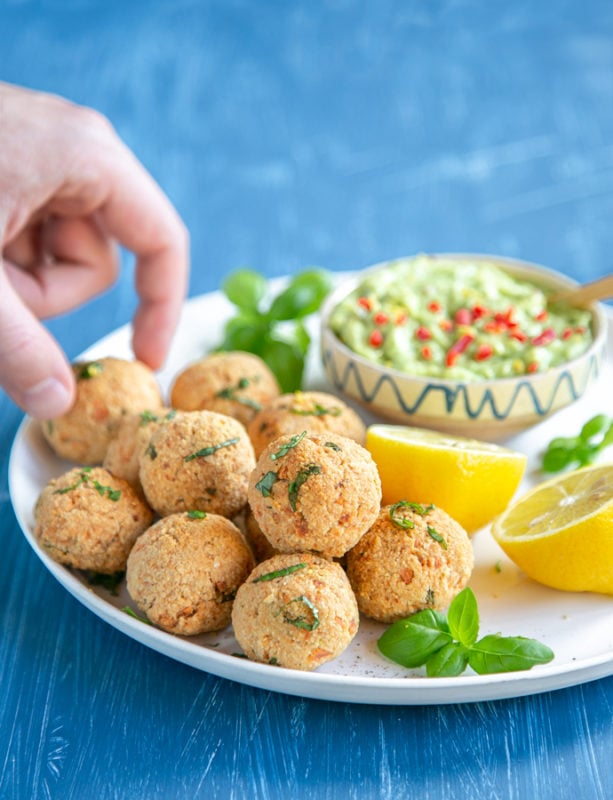baked salmon meatballs on white plate served with halved lemon, basil leaves and avocado cream in a small pot, hand picking up a meatball in the background